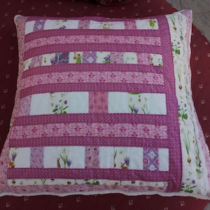 Op de afbeelding: Een roze en wit patchwork kussen met een bloemenpatroon. Het kussen is gemaakt van verschillende stoffen en heeft een witte rand.