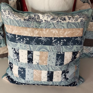 Könnte beinhalten: Ein quadratisches Zierkissen mit Patchwork-Design. Das Kissen zeigt verschiedene blaue und beige Blumen- und Paisley-Muster. Das Kissen hat einen hellblauen Rand und wird wahrscheinlich für die Inneneinrichtung verwendet.