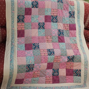 Könnte beinhalten: Ein Patchwork-Quilt mit verschiedenen gemusterten Quadraten in Rosa-, Türkis- und Lilatönen. Der Quilt hat einen weißen Rand mit einer türkisfarbenen und rosa gemusterten Bordüre. Der Quilt ist über einer rot gemusterten Oberfläche drapiert.