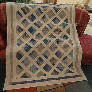 Peut inclure: Une courtepointe rectangulaire présentant un motif en treillis de bandes de tissu crème croisant des carrés de tissus aux motifs floraux et géométriques variés dans des tons de bleu, de beige et de brun, avec une bordure florale décorative.