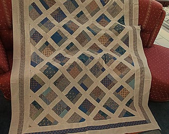 Può includere: Una trapunta rettangolare caratterizzata da un motivo a reticolo di strisce di tessuto color crema che si incrociano su toppe quadrate di vari tessuti con motivi floreali e geometrici blu, marroni e color cuoio, con un bordo floreale.