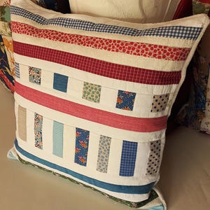 Peut inclure: Un oreiller blanc avec un motif patchwork composé de rayures et de carrés rouges, bleus et roses. L'oreiller est fait de différents tissus avec divers motifs, notamment des pois, du vichy et des imprimés floraux.