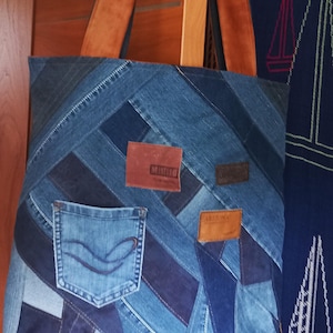 Op de afbeelding: Een blauwe denim tote bag gemaakt van gerecyclede jeans. De tas heeft een patchwork ontwerp met verschillende tinten blauw denim en bruine leren patches. De patches hebben de woorden "Mister" en "Arizona" erop gedrukt.