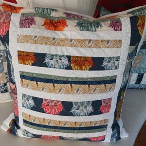 Puede incluir: Una almohada blanca con una tela estampada azul y blanca. La almohada tiene un borde blanco y está decorada con rayas de colores y diseños florales.