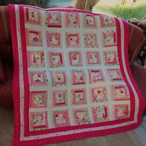 Op de afbeelding: Een roze en witte patchworkdeken met een roze rand en een witte achtergrond. De deken is gemaakt van vierkanten met verschillende ontwerpen, waaronder feeën, bloemen en andere fantasierijke patronen.