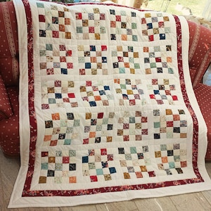 Può includere: Una trapunta fatta a mano con sfondo bianco e design patchwork. La trapunta presenta una griglia di piccoli quadrati in vari colori, tra cui rosso, blu e marrone. Il bordo è rosso scuro con un motivo floreale.