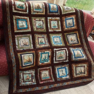 Könnte beinhalten: Eine braune Quiltdecke mit nautischem Thema, die quadratische Paneele mit Bildern von Segelbooten, Leuchttürmen und Seekarten zeigt. Die Decke hat einen dunkelbraunen Rand und wird auf einem roten Sofa präsentiert. Die Quadrate sind mit verschiedenen Farben umrahmt.