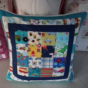 Könnte beinhalten: Ein blaues und weißes Patchwork-Kissen mit verschiedenen bunten Stoffquadraten mit Autos, Tieren und anderen Designs. Das Kissen hat einen blauen Rand und einen weißen Hintergrund.