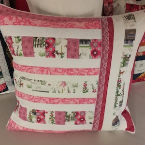 Può includere: Un cuscino bianco con tessuto a righe rosa e bianco e un motivo floreale rosa e bianco. Il cuscino ha un bordo in pizzo bianco e un bordo rosa.