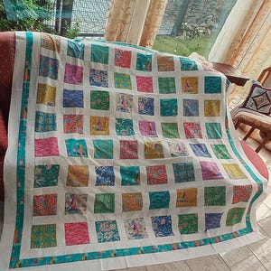 Peut inclure: Un quilt en patchwork composé d'une grille de carrés de tissu colorés dans des tons de bleu sarcelle, rose, jaune et vert, séparés par des bandes blanches. Le quilt présente une bordure bleu sarcelle et divers motifs floraux sur chaque carré.