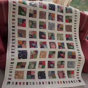 Könnte beinhalten: Eine weiße Steppdecke mit Patchwork-Muster, das aus Quadraten aus verschiedenen Blumenstoffen besteht. Die Steppdecke hat einen weißen Rand mit einem sich wiederholenden Muster aus kleinen Quadraten in verschiedenen Farben.