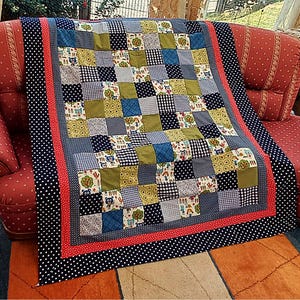 Könnte beinhalten: Eine Patchwork-Decke mit einem Raster aus Quadraten in verschiedenen Mustern, darunter blaues Vichy-Karo, Olivgrün, marineblaue Punkte sowie Drucke von Zeichentricktieren und Bäumen. Die Decke ist mit einem roten Band und einem breiten Rand eingefasst.