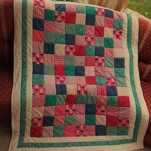 Op de afbeelding: Een kleurrijke patchworkquilt met een vierkant patroon in roze, turquoise, rood en blauw. De quilt heeft een witte rand met turquoise strepen. De quilt is over een rode bank gedrapeerd.