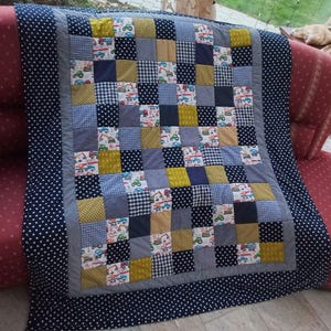Könnte beinhalten: Eine blaue und gelbe Patchwork-Decke mit verschiedenen Mustern, darunter Punkte, Gingham und ein Druck mit Baufahrzeugen. Die Decke besteht aus Quadraten und ist mit einem dunkelblauen Stoff mit weißen Punkten eingefasst.