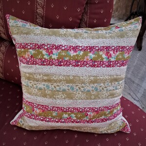 Puede incluir: Una almohada decorativa con un diseño de patchwork que presenta rayas de tela rosa, blanca y verde con patrones florales. La almohada está hecha de tela acolchada y tiene una textura suave y cómoda.