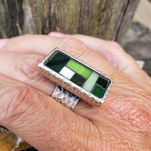May include: A rectangular silver ring with a mosaic design. The mosaic features a pattern of green, white, and light green tiles. The ring has a hammered texture on the band and a bezel setting.