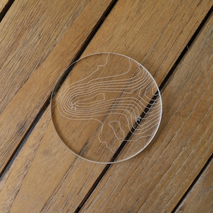 May include: A clear round coaster with a topographic map design etched into the surface. The coaster is on a wooden surface.