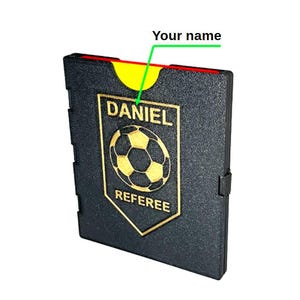 May include: A black referee wallet featuring a gold-coloured emblem with a football graphic, the text DANIEL, and the word REFEREE. The wallet includes a yellow card slot at the top, with a green arrow pointing to the name area labelled Your name.