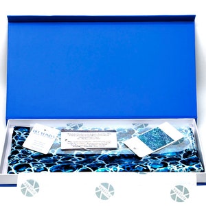 May include: A blue box with a white interior, containing a blue and white patterned silk scarf. The scarf is folded and has a small tag attached to it. The tag says "Blomonda".