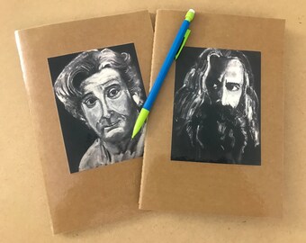 May include: Two brown notebooks with black and white portraits on the covers. A blue and green pencil rests between the notebooks. The portraits are of two men with long hair and beards.