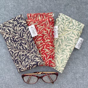May include: Three fabric eyeglass cases with a botanical leaf pattern in navy, red, and cream. The cases are rectangular and have a tag that says "Stitched Up!" A pair of eyeglasses with a red and brown frame rests below the cases.