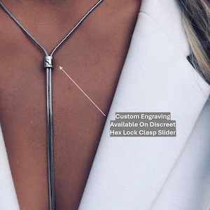 Può includere: Una collana argentata con un pendente a catena verticale. La collana presenta una chiusura scorrevole e il testo "Custom Engraving Available On Discreet Hex Lock Clasp Slider". La collana è indossata sopra un blazer bianco.