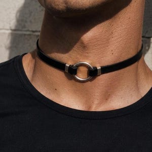 Könnte beinhalten: Ein schwarzes Lederhalsband mit einem zentralen Ring aus silberfarbenem Metall. Das Band ist auf beiden Seiten des Rings mit silberfarbenen Metallbeschlägen befestigt, wodurch ein kreisförmiger Blickfang am Halsbereich entsteht.