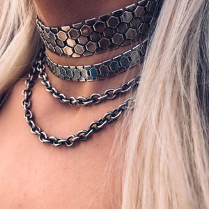 Può includere: Un set di collane a strati composto da due ampie fasce girocollo piatte con un motivo metallico esagonale ripetuto e due catene a maglie color argento di diverse lunghezze che ricadono a cascata sotto i girocollo in una disposizione a livelli.