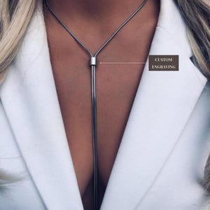 May include: A silver necklace with a vertical chain pendant. The necklace is worn over a white blazer. The text "CUSTOM ENGRAVING" is visible in a small brown box.
