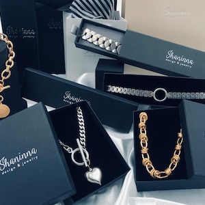 May include: A collection of gold and silver necklaces and bracelets displayed in black boxes with the logo "Shaninna Design & Jewelry".