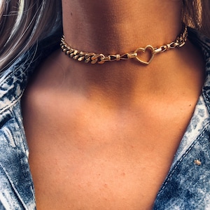 Könnte beinhalten: Goldfarbene Choker-Halskette mit einem Panzerketten-Design. Die Halskette hat einen herzförmigen Anhänger in der Mitte. Der Schmuck wird am Hals einer Person getragen, vor einer Jeansjacke.