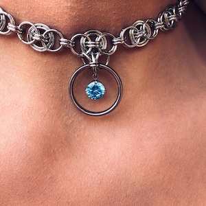 May include: A silver-toned chainmail choker necklace constructed from interlocking metal rings. The center of the necklace features a circular metal pendant holding a single, round, faceted bright blue gemstone suspended in the middle.