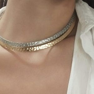 Könnte beinhalten: Eine mehrlagige Choker-Halskette in Silber und Gold. Die Halskette besteht aus zwei dünnen Metallbändern, einem silbernen und einem goldenen. Das silberne Band ist strukturiert und das goldene Band ist glatt.