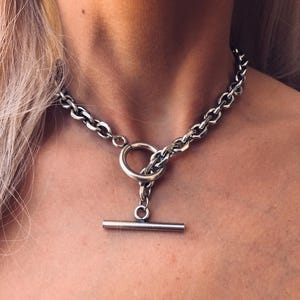Op de afbeelding: Een zilveren ketting met een T-vormige sluiting. De ketting is dik en heeft een touwachtige textuur.