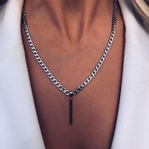 Puede incluir: Un collar de cadena plateada con un colgante rectangular. El collar es de cadena de eslabones y cuelga sobre una blazer blanca. El colgante es una barra vertical simple. Es una joya.