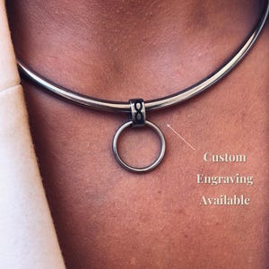 May include: A dark metallic choker necklace featuring a smooth, rigid band and a central circular pendant. The pendant is attached to the band by a small rectangular connector engraved with an infinity symbol. The text Custom Engraving Available is visible.