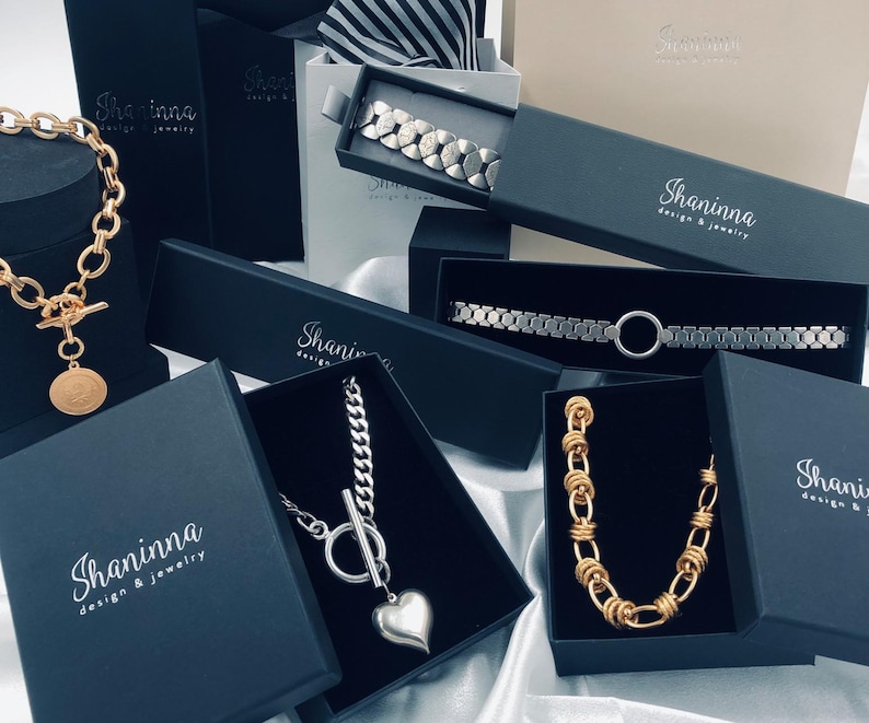 May include: A collection of gold and silver necklaces and bracelets displayed in black boxes with the logo "Shaninna Design & Jewelry".