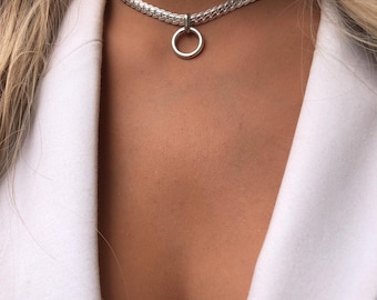 Pode incluir: Um colar gargantilha prateado com um grande pendente circular. O colar tem um design de corrente grossa e tecida. O pendente é um anel simples e polido. O fundo inclui um blazer branco e cabelo loiro.