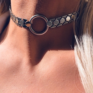 May include: A silver-toned metal choker necklace featuring a series of interconnected hexagonal and triangular links. The design centres on a smooth, circular ring element that connects the two sides of the metallic chain, resting against the neck.
