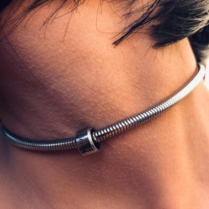 Peut inclure: Un collier ras du cou argenté avec un motif de chaîne serpent. Le collier comporte un petit charme cylindrique avec du texte gravé. Le bijou est porté autour du cou.