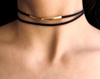 May include: A black choker necklace with a gold bar pendant. The necklace is made of two layers of thin black cord.