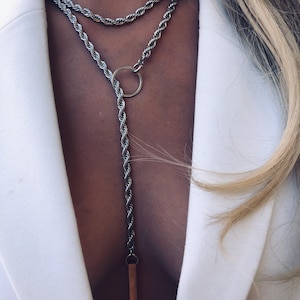 May include: Silver-toned rope chain necklace with a layered design. The necklace features a circular pendant and a long, dangling element with a rectangular charm. The jewelry is displayed against a white blazer.