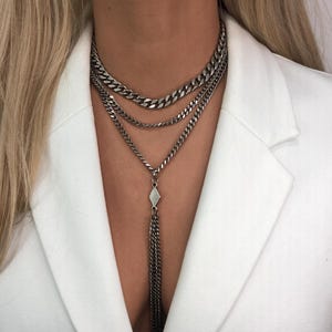 May include: A multi-layered silver-toned chain necklace featuring three distinct curb chain strands of varying lengths. The longest strand connects to a central diamond-shaped metal charm, which suspends a long, thin, dangling chain tassel.