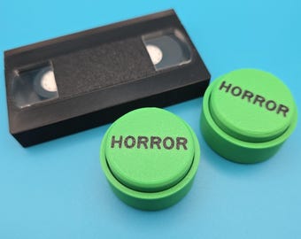 May include: Two green buttons with the word "HORROR" printed on them, next to a black VHS tape.