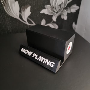 May include: A black, rectangular phone stand with a white "NOW PLAYING" sign. The stand has a circular design with a red center and white border. The stand is on a black surface.