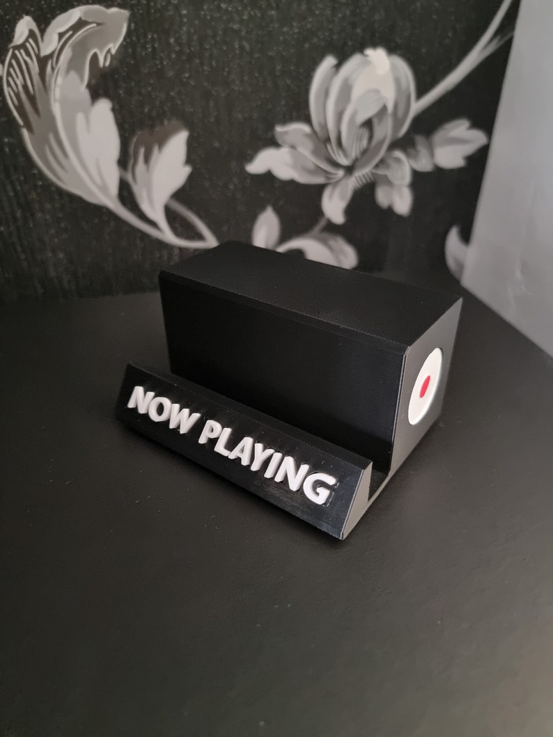 May include: A black, rectangular phone stand with a white "NOW PLAYING" sign. The stand has a circular design with a red center and white border. The stand is on a black surface.