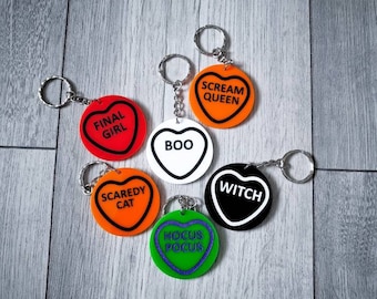May include: Six heart-shaped keychains with Halloween-themed sayings. The keychains are orange, red, green, white, and black. The sayings include "Final Girl", "Boo", "Scaredy Cat", "Hocus Pocus", "Witch", and "Scream Queen".