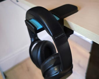 May include: Black over-ear headphones hanging from a black desk clamp with a light blue accent. The headphones are labeled "WH-1000XM3". The clamp is attached to a light-colored wooden desk.
