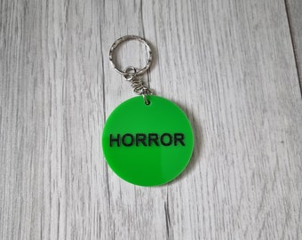 May include: Green round keychain with a silver key ring and the word "HORROR" printed in black.
