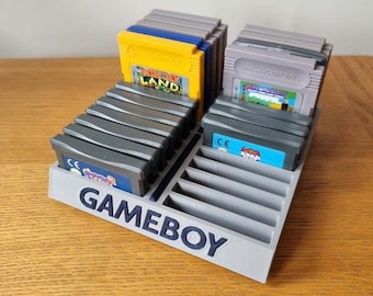 May include: A gray plastic Game Boy cartridge holder with a blue label that says "GAMEBOY". The holder has 16 slots for cartridges. There are 14 gray Game Boy cartridges in the holder, and two yellow Game Boy cartridges. One of the yellow cartridges has the text "DONKEY KONG LAND" on it. The other yellow cartridge has the text "SUPER MARIO LAND 2: 6 GOLDEN COINS" on it.